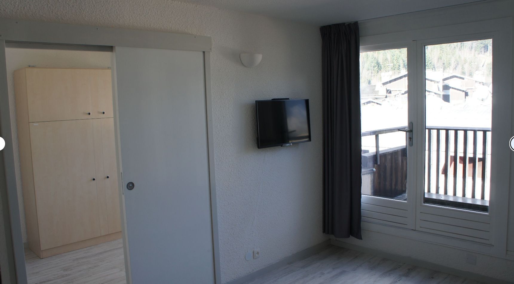 Una stanza con una porta chiusa, una televisione a parete e vista su un balcone esterno con ringhiera.