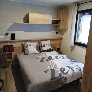 A bedroom with a bed, bedside tables, a wooden headboard, a window, and a wall-mounted shelf. - Rental of a fully equipped 40-square-meter mobile home with a 20-square-meter covered patio (La Tremblade)