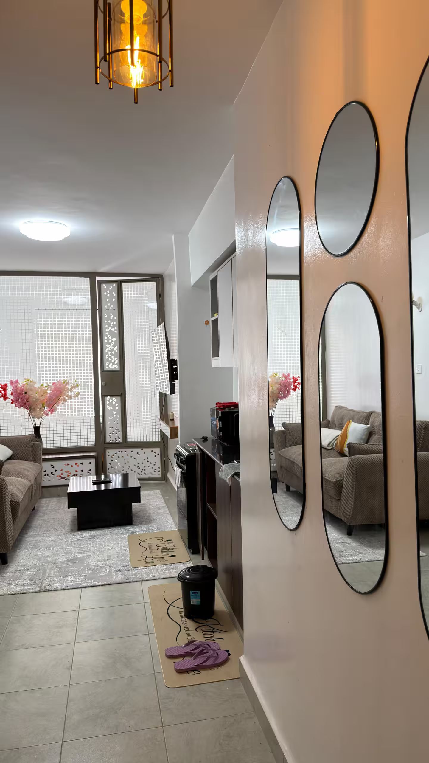 Een moderne woonkamer met een bank, een salontafel en een televisie. Er hangen drie spiegels aan de muur en er is een plafondlamp.