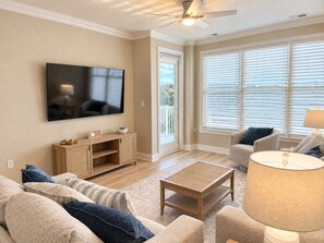 A living room with a flat-screen TV, a wooden entertainment unit, a sofa set, a coffee table, and a lamp. - Luxury Beach Condo | Pool | Surf | Swim | Arcade (Virginia Beach)