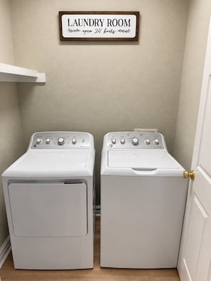 A laundry room with two white front-loading washing machines and a sign above them stating 'LAUNDRY ROOM open 24 hours.' - Luxury Beach Condo | Pool | Surf | Swim | Arcade (Virginia Beach)