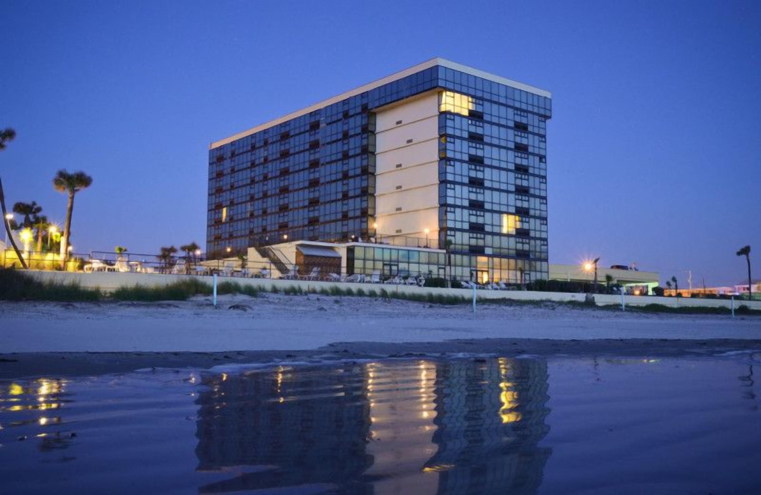 Un hôtel moderne à la façade en verre réfléchissant, situé au bord d’une plage dont le sable mouillé s’étend jusqu’au rivage.