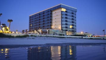 Un hôtel moderne à la façade en verre réfléchissant, situé au bord d’une plage dont le sable mouillé s’étend jusqu’au rivage.