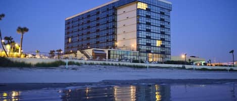 Un hotel moderno con fachada de vidrio reflectante, ubicado junto a una playa con arena húmeda en primer plano.