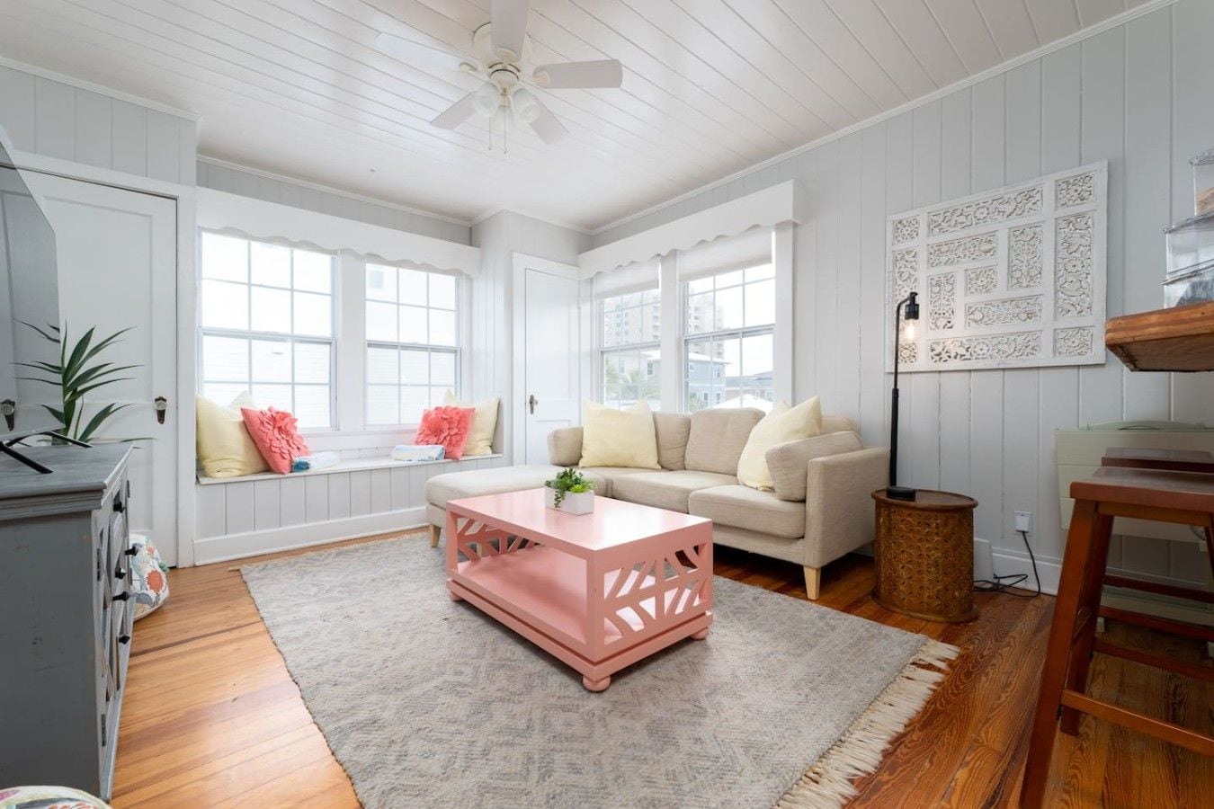 A room with a white ceiling, a ceiling fan, a window seat with cushions, a sofa, a pink ottoman, a rug, a wooden floor, and a wall with decorative panels.