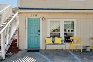 A building with a teal door, a number '126' above it, a bench with yellow cushions, and a yellow chair outside. - Turning Tide · 1BR · No Stairs · Pet friendly (Jacksonville Beach)