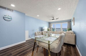 A room with a glass dining table, beige upholstered chairs, a ceiling fan, and a view through a window. - The Allura- Galveston Condo Steps Away from Beach. Perfect for 4 people! (Galveston)
