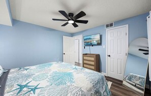 Ein Schlafzimmer mit einem Bett, einem Deckenventilator, einem Schminktisch, einem Fernseher und einer Tür.