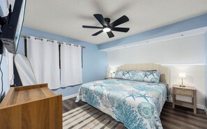 A bedroom with a bed, curtains, a ceiling fan, a bedside table, and a nightstand. - The Allura- Galveston Condo Steps Away from Beach. Perfect for 4 people! (Galveston)