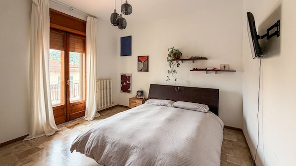 A bedroom with a bed, a radiator, a window with curtains, and wall-mounted shelves. - defaultName (Brienza)