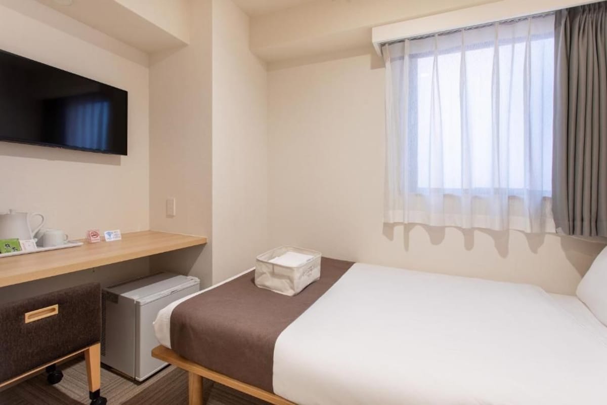 Een hotelkamer met een bed, een bureau met een flatscreen televisie, een minibar en een raam met dunne gordijnen.