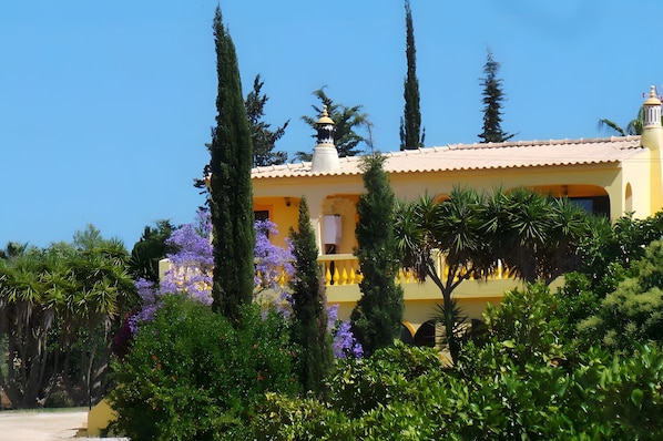 A yellow building with a tiled roof, surrounded by greenery and tall cypress trees.