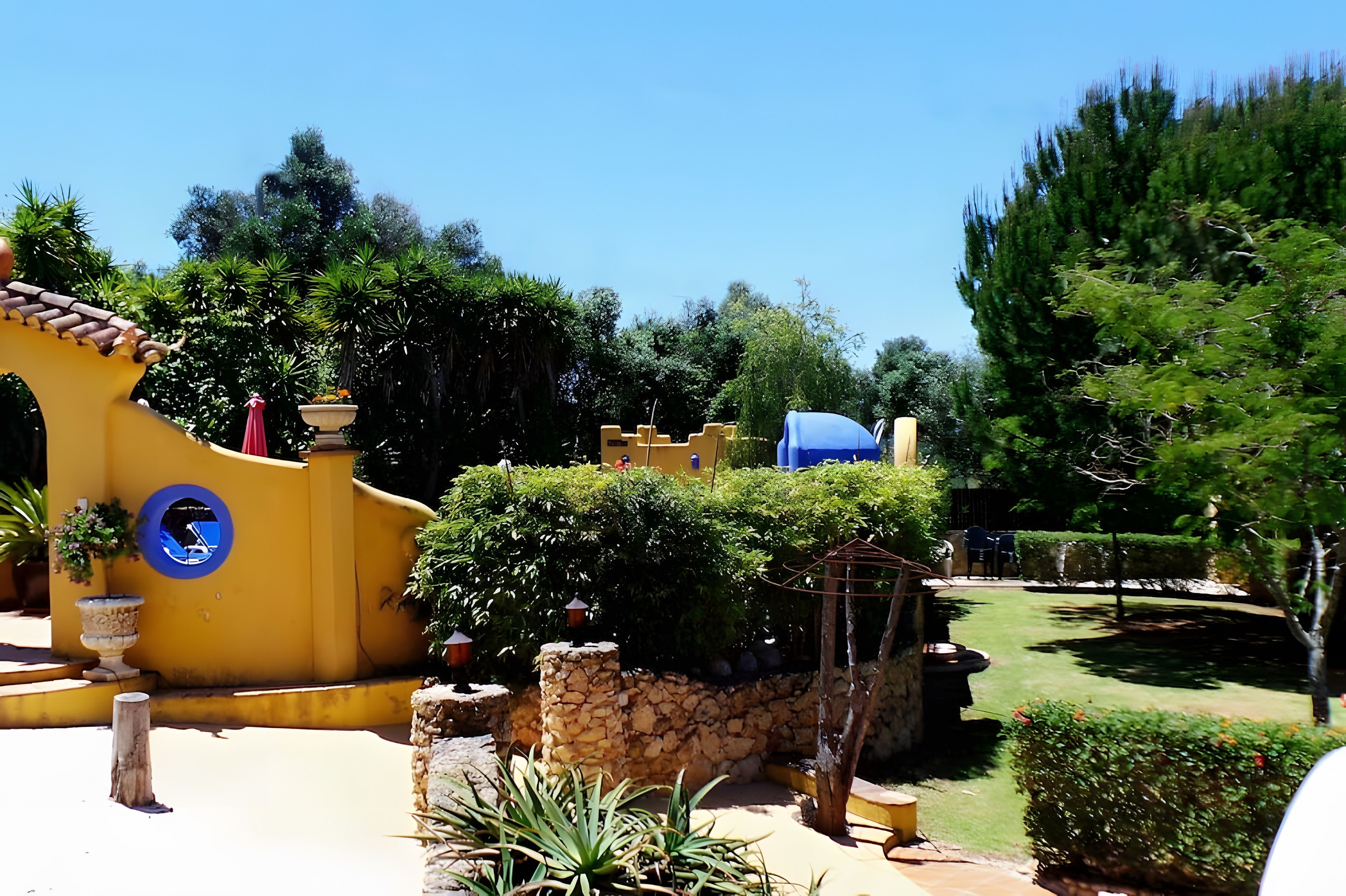 Un jardín con pared amarilla, estructuras de piedra y diversas plantas.