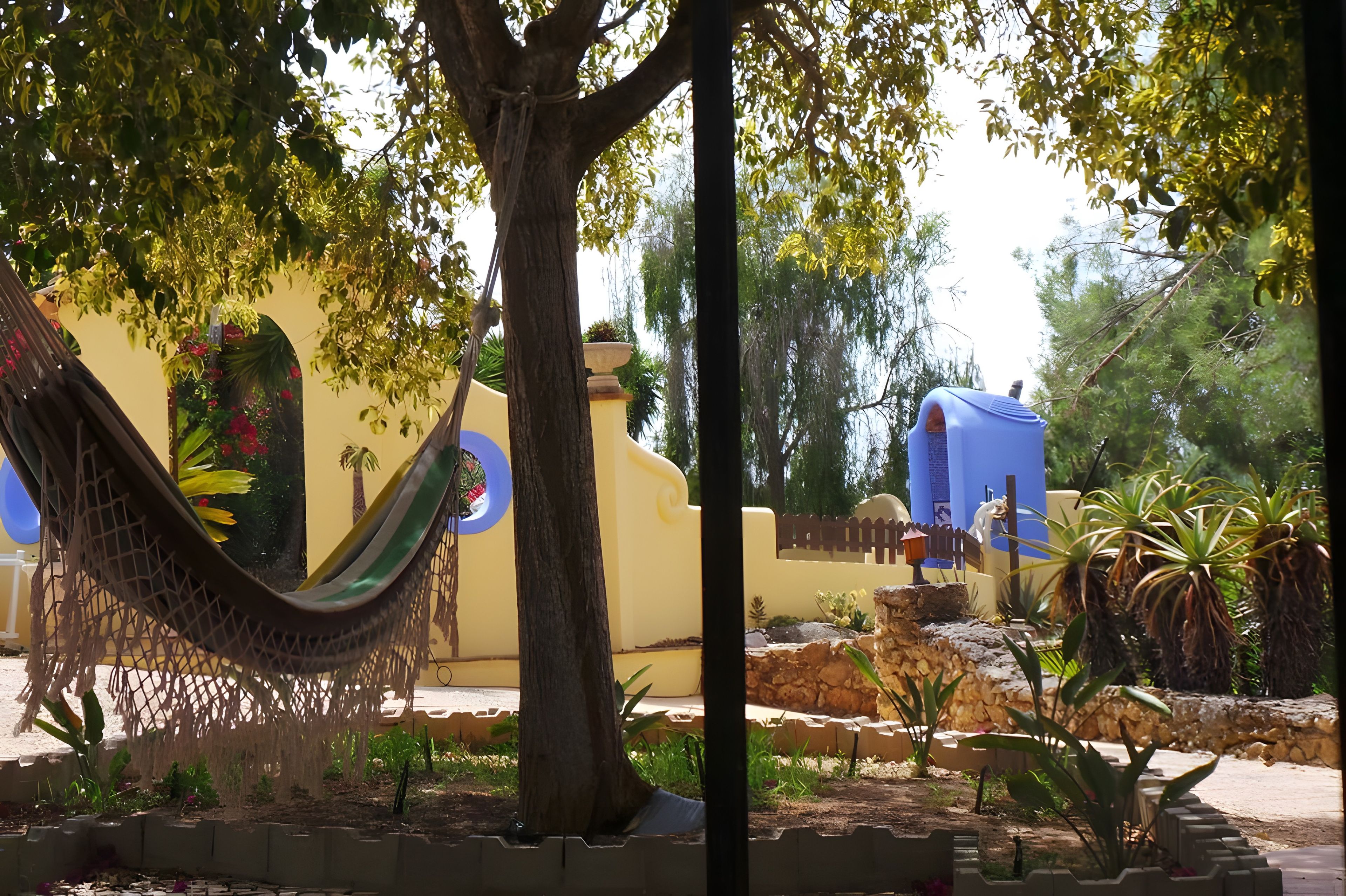 A hammock hanging between trees, a yellow wall with a mural, and a blue structure in a garden setting.