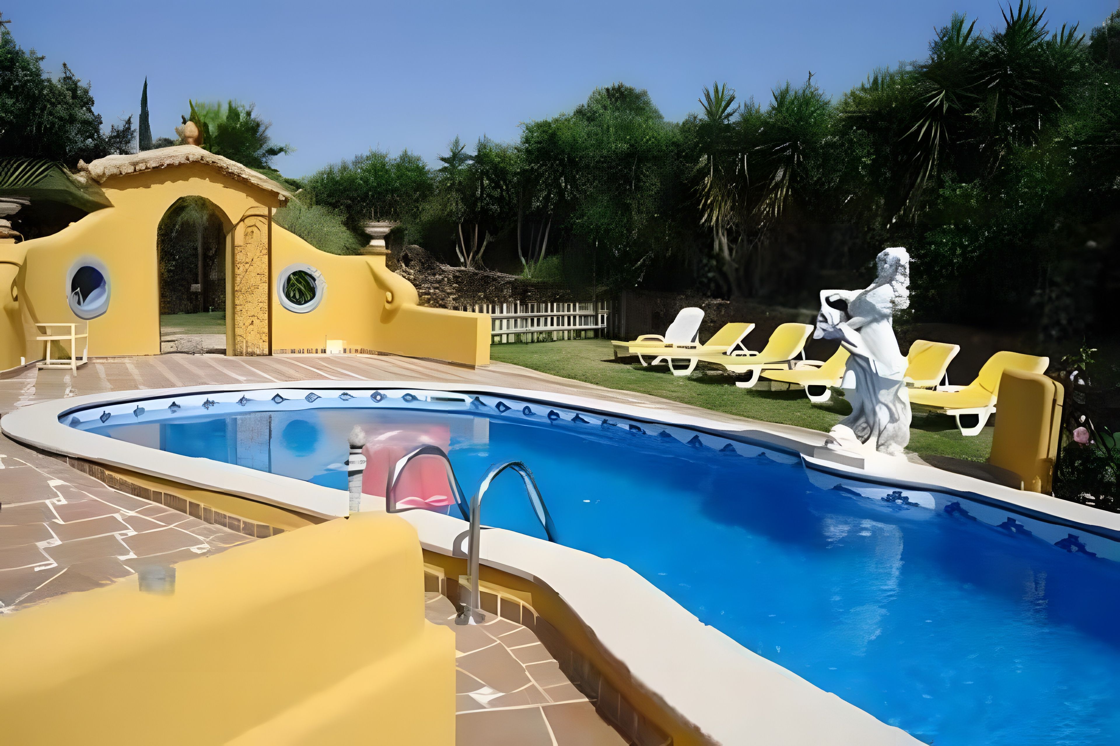 A swimming pool with a statue, yellow lounge chairs, and a small building with a circular window.