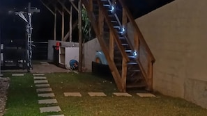 Un espace extérieur nocturne comprenant un escalier en bois, une voiture bleue et une structure blanche.