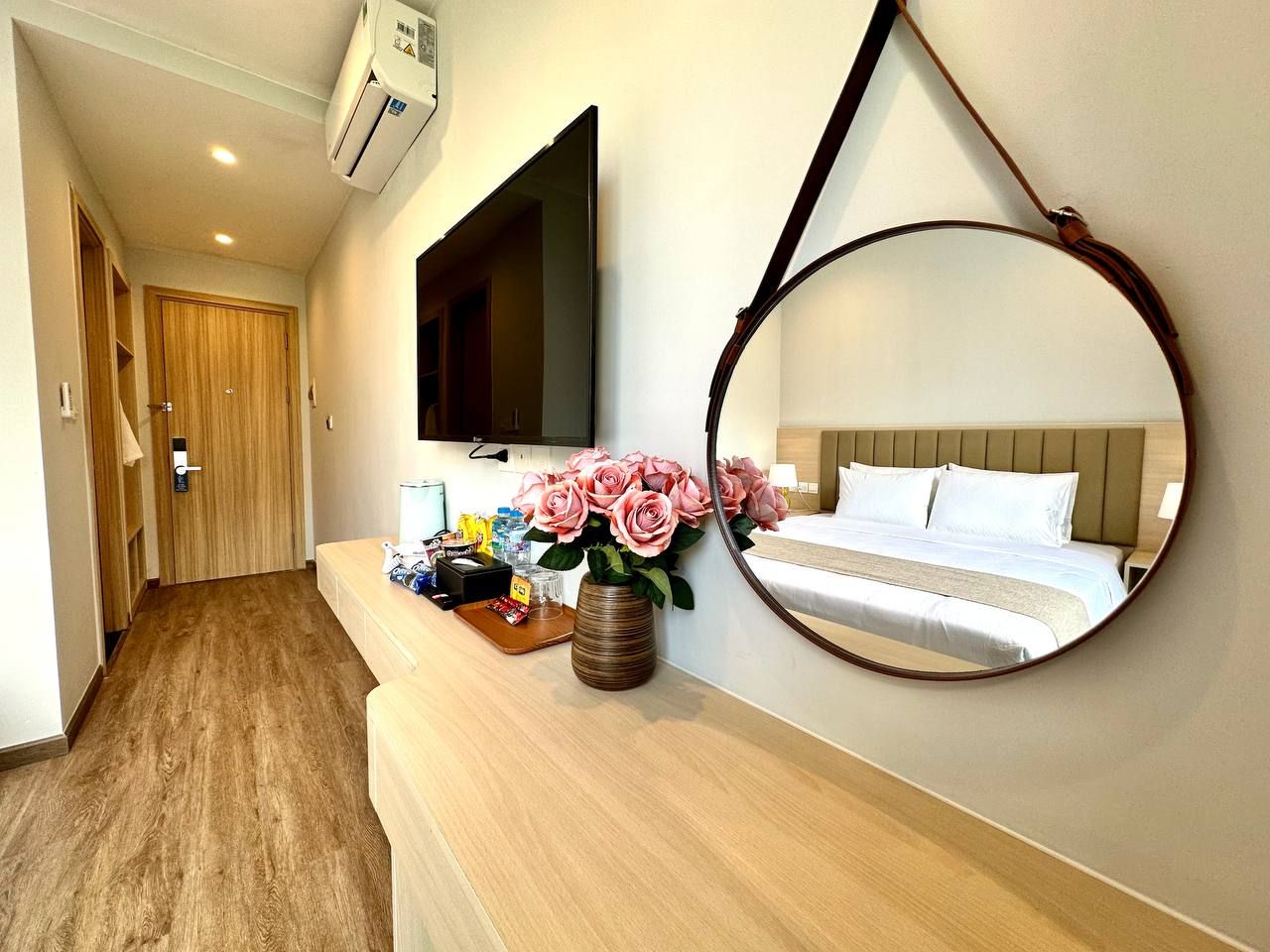A hotel room with a bed, a TV mounted on the wall, a mirror, and a vase with flowers on the bedside table.