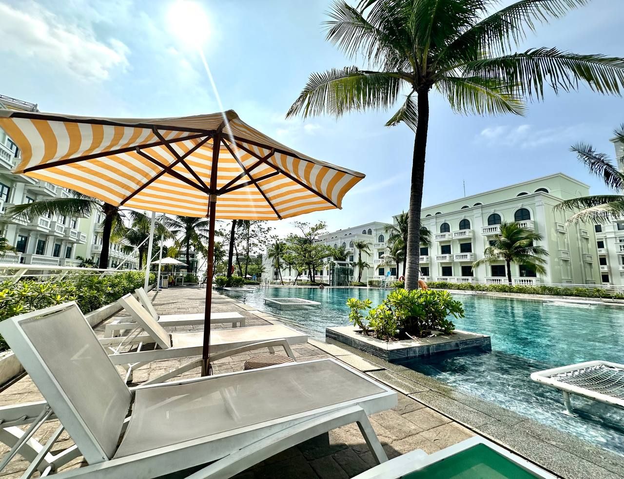 A poolside area with lounge chairs and a large striped umbrella, surrounded by palm trees and a building with multiple balconies.