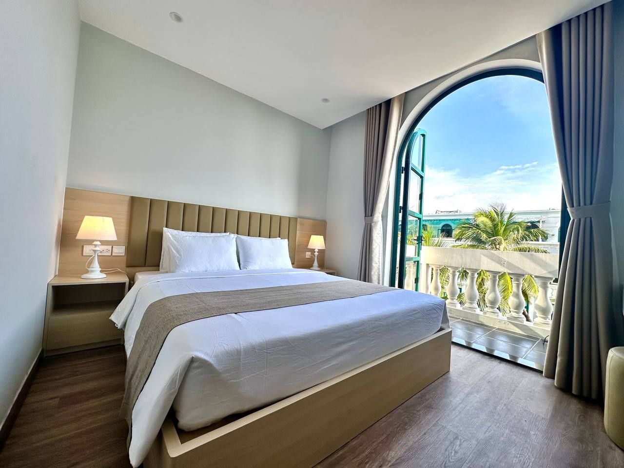 A hotel room with a large bed, a nightstand, a window with curtains, and a view of palm trees.
