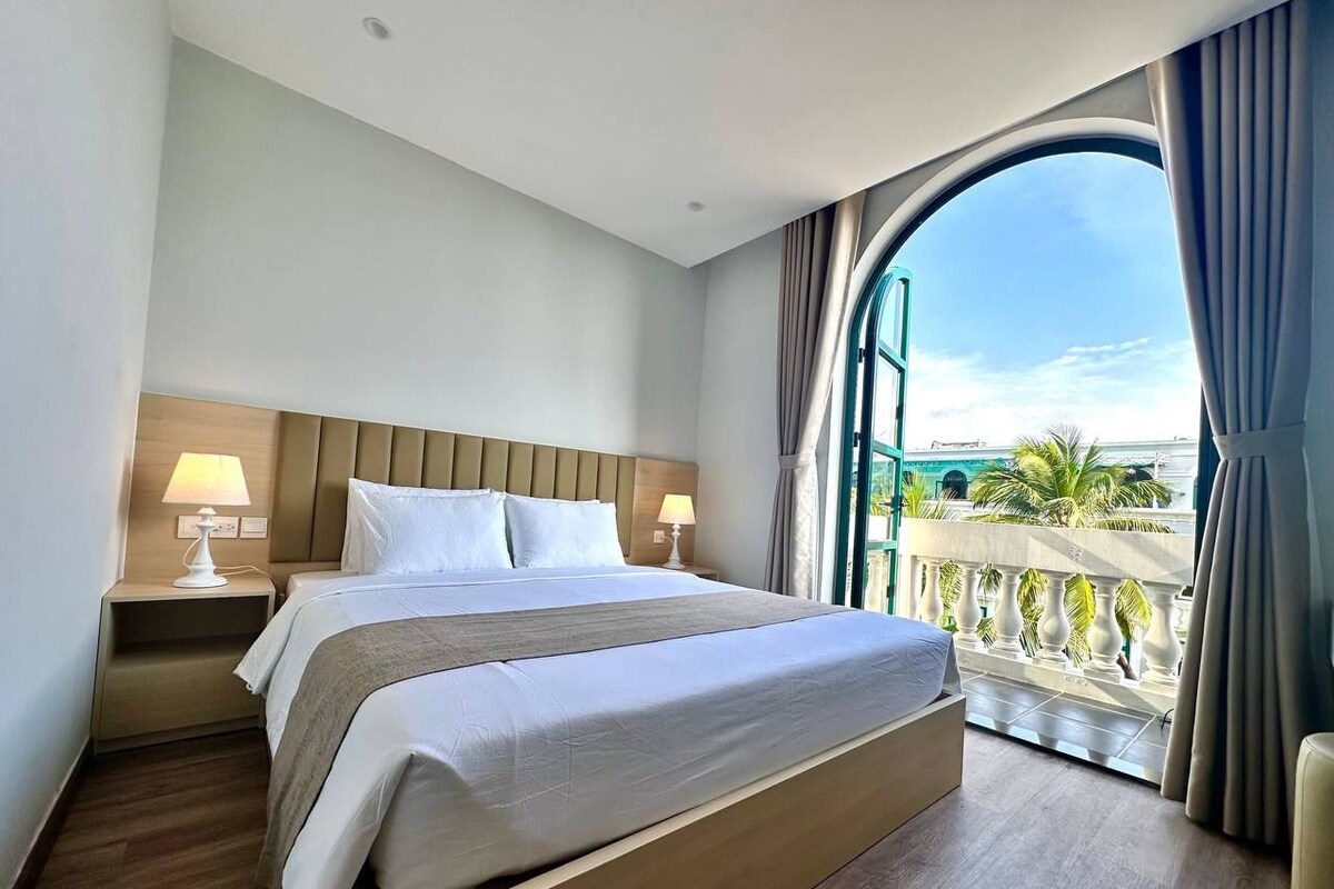Quarto de hotel com uma cama grande, mesa de cabeceira, janela com cortinas e vista para palmeiras.