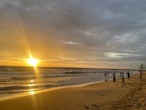 Escena de playa con el sol poniéndose sobre el océano, gente en la orilla y un barco en el agua.