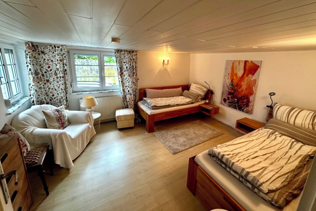 Ein Schlafzimmer mit Bett, Stuhl, Nachttisch, Lampe, Heizkörper und einem Fenster mit Vorhängen.