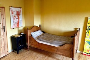Un simple lit en bois avec un matelas et des oreillers, une petite table de chevet noire sur laquelle se trouve un livre, et une porte sur la gauche.