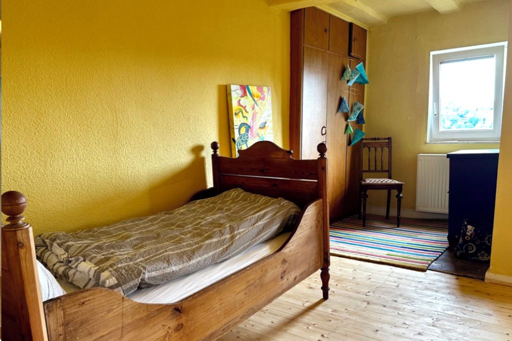 Un lit simple avec un cadre en bois, dans une pièce aux murs jaunes et dotée d’une fenêtre.