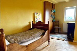 Un lit simple avec un cadre en bois, dans une pièce aux murs jaunes et dotée d’une fenêtre.