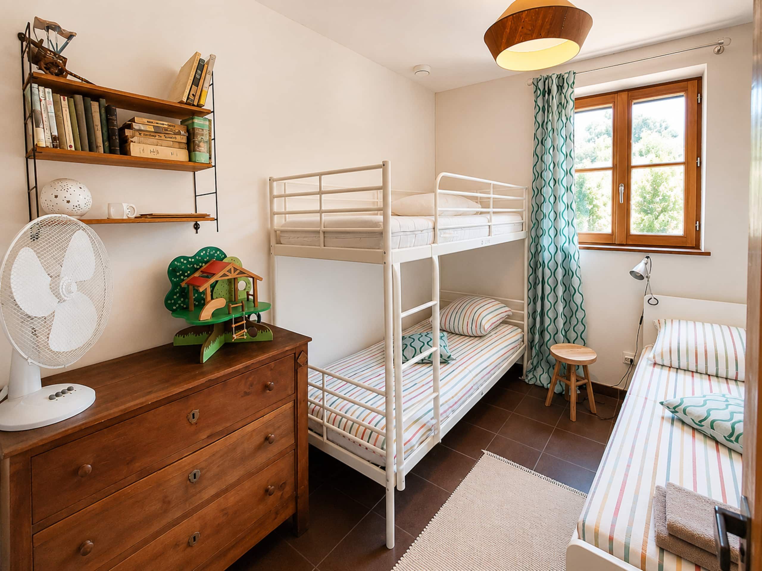 Une chambre avec un lit superposé, une commode en bois, un ventilateur, une bibliothèque et une fenêtre avec des rideaux.