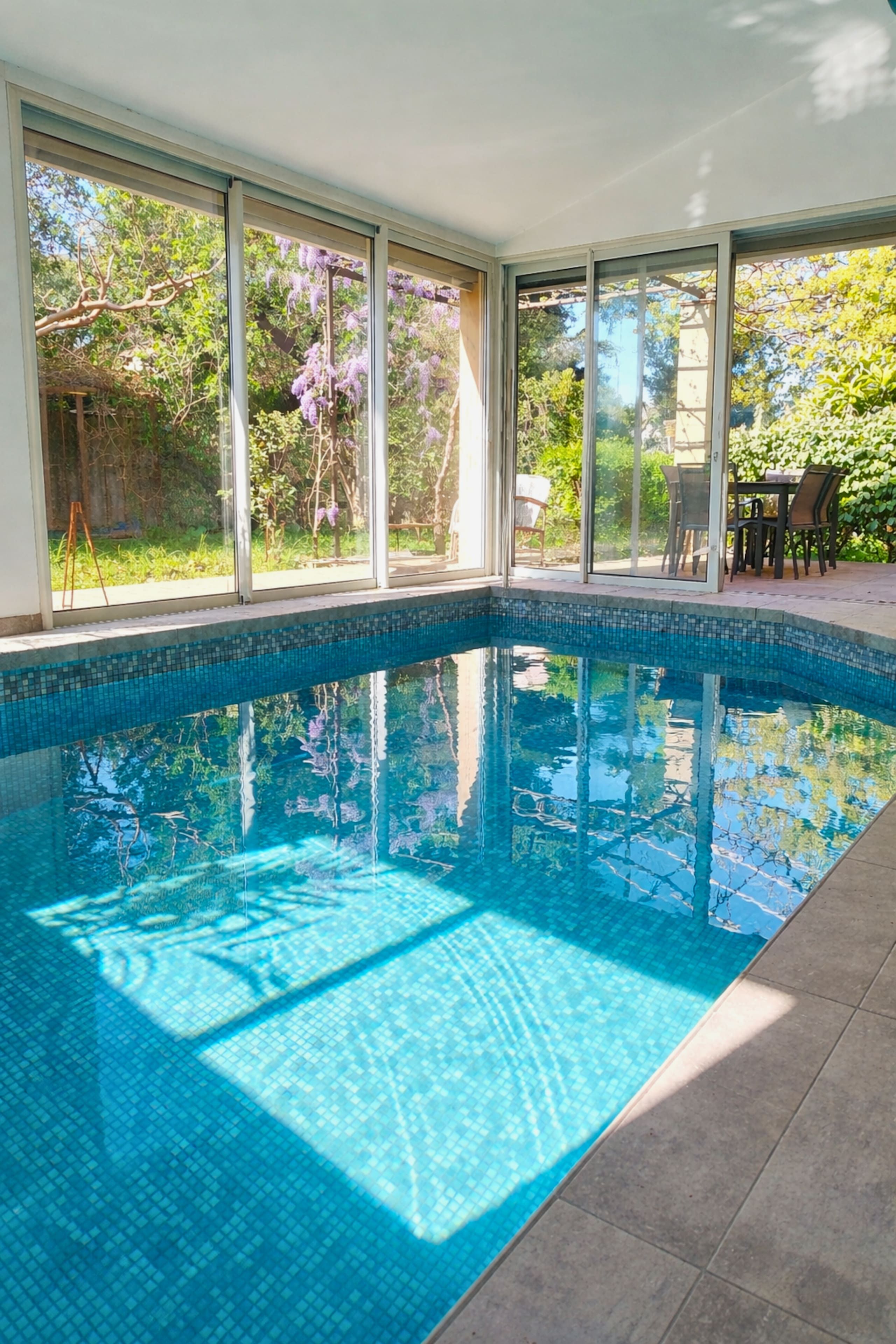 A clear indoor swimming pool with a tiled edge, surrounded by large windows offering views of greenery.
