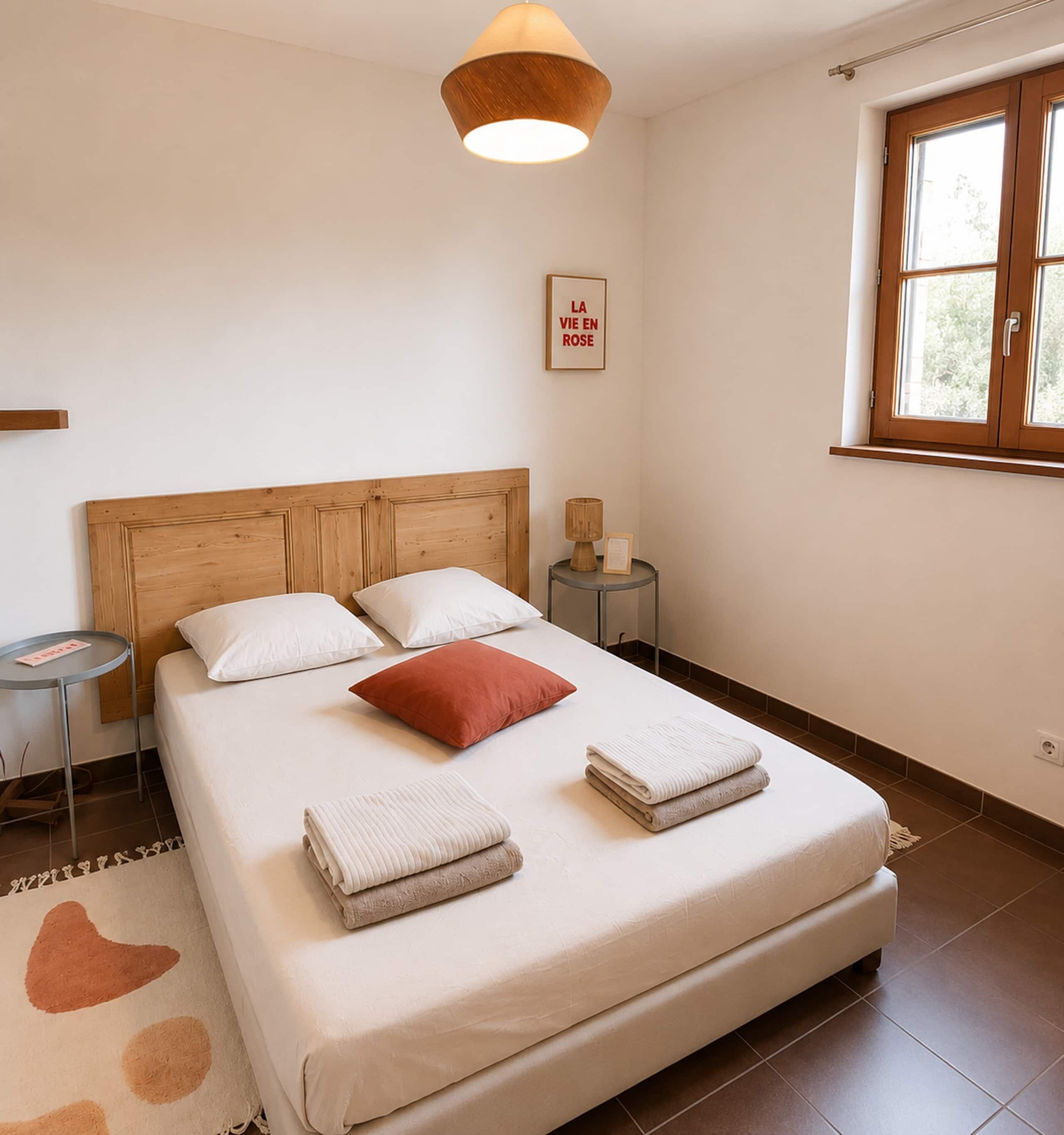 Ein Schlafzimmer mit hölzernem Kopfende, einem Bett mit weiß-roten Kissen, einem Nachttisch mit Lampe und einem Fenster mit Blick.