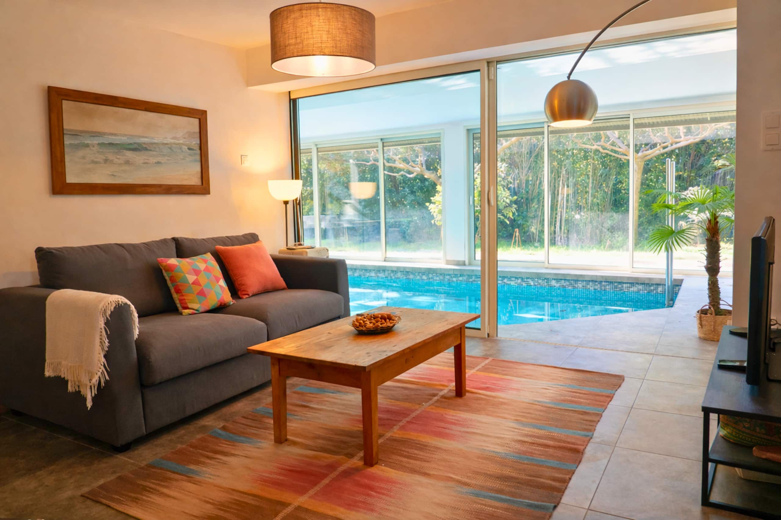 Un soggiorno con un divano grigio, cuscini colorati, un tavolino in legno e una grande finestra vista piscina.