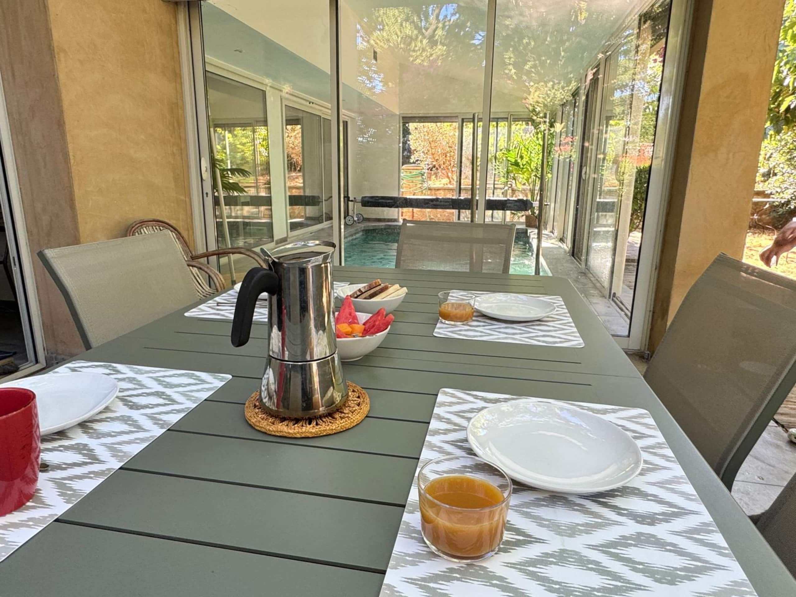A dining table with a metal coffee pot, a glass of juice, and a bowl of fruit.