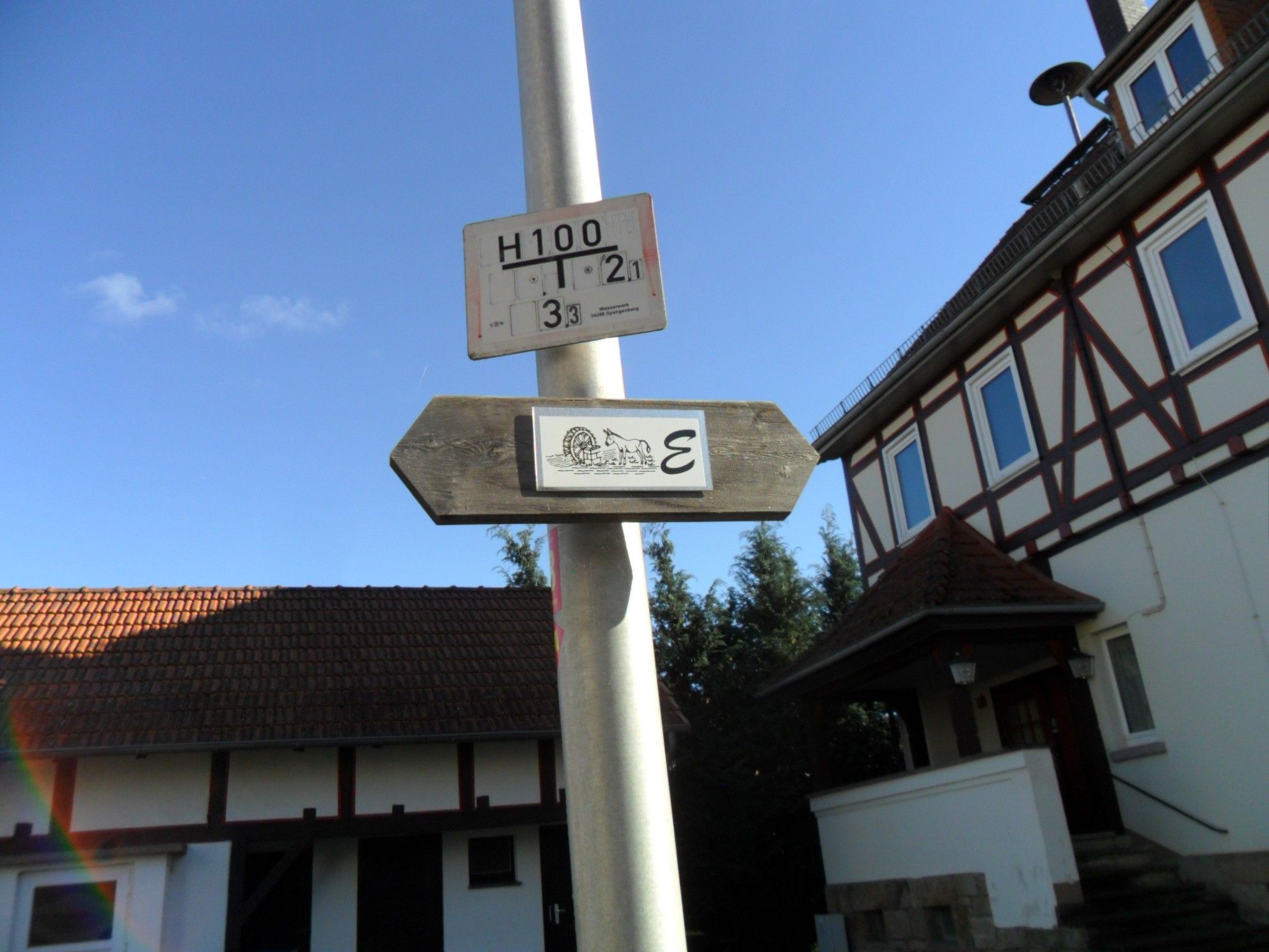 Ein Wegweiser mit mehreren Schildern, darunter eines mit der Aufschrift 'H100' und ein weiteres mit einem Symbol und dem Buchstaben 'E'.