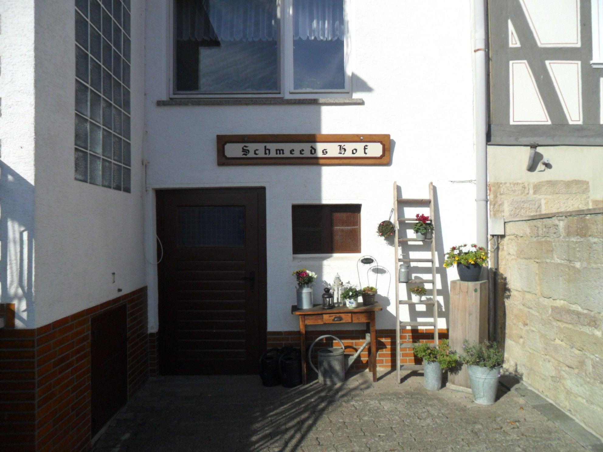 A building with a wooden door and a sign that reads 'Schmeeds Hoef'. There is a wooden bench with potted plants beside it, and a ladder leaning against the wall.