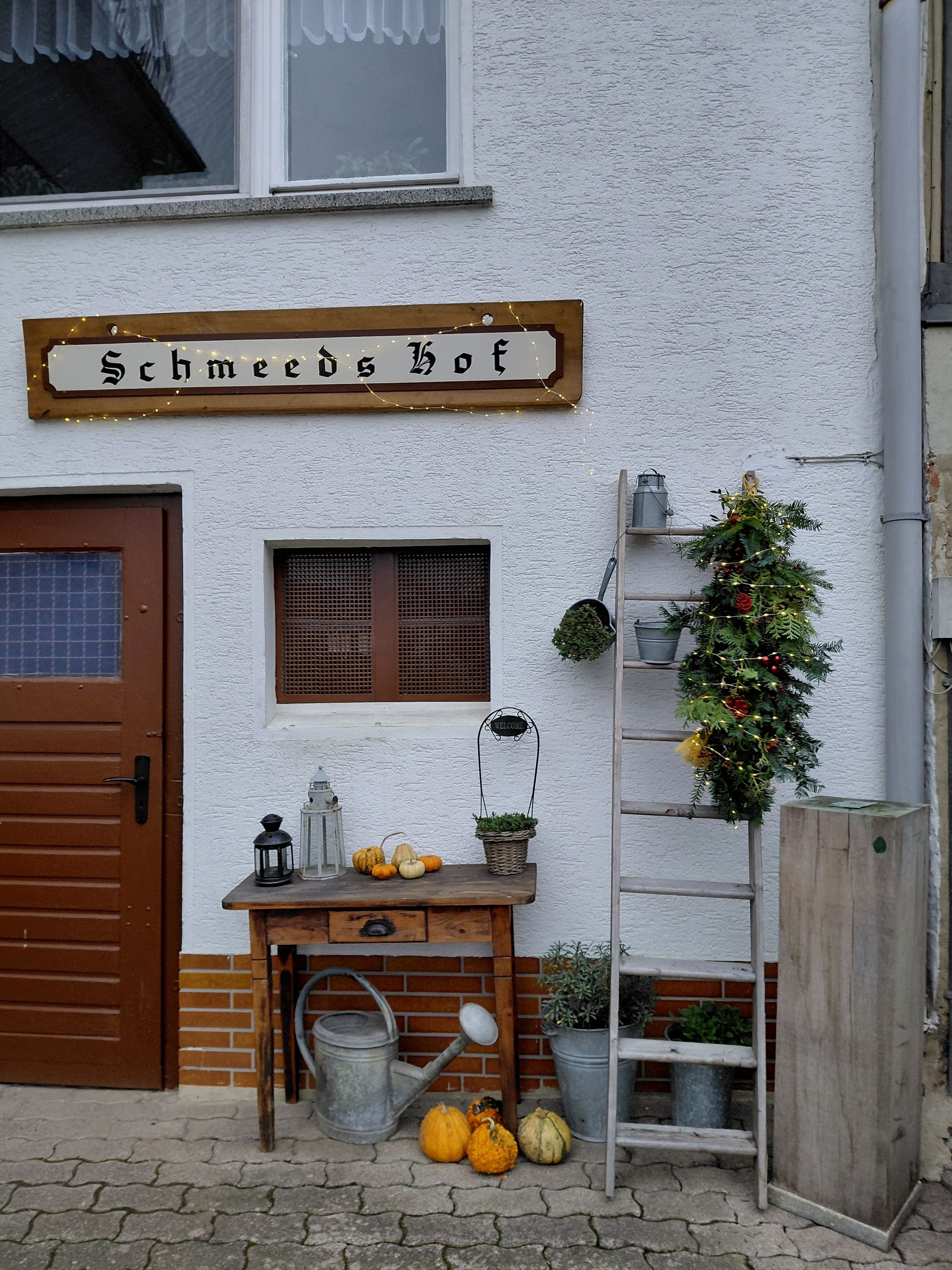 Een houten bord met de naam 'Schmeeds Hof' boven een deur, een klein raam met luiken, een welkomstbord, een kerstboom en een houten bank met pompoenen en een lantaarn.