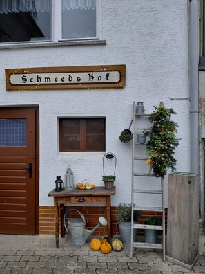 Een houten bord met de naam 'Schmeeds Hof' boven een deur, een klein raam met luiken, een welkomstbord, een kerstboom en een houten bank met pompoenen en een lantaarn.