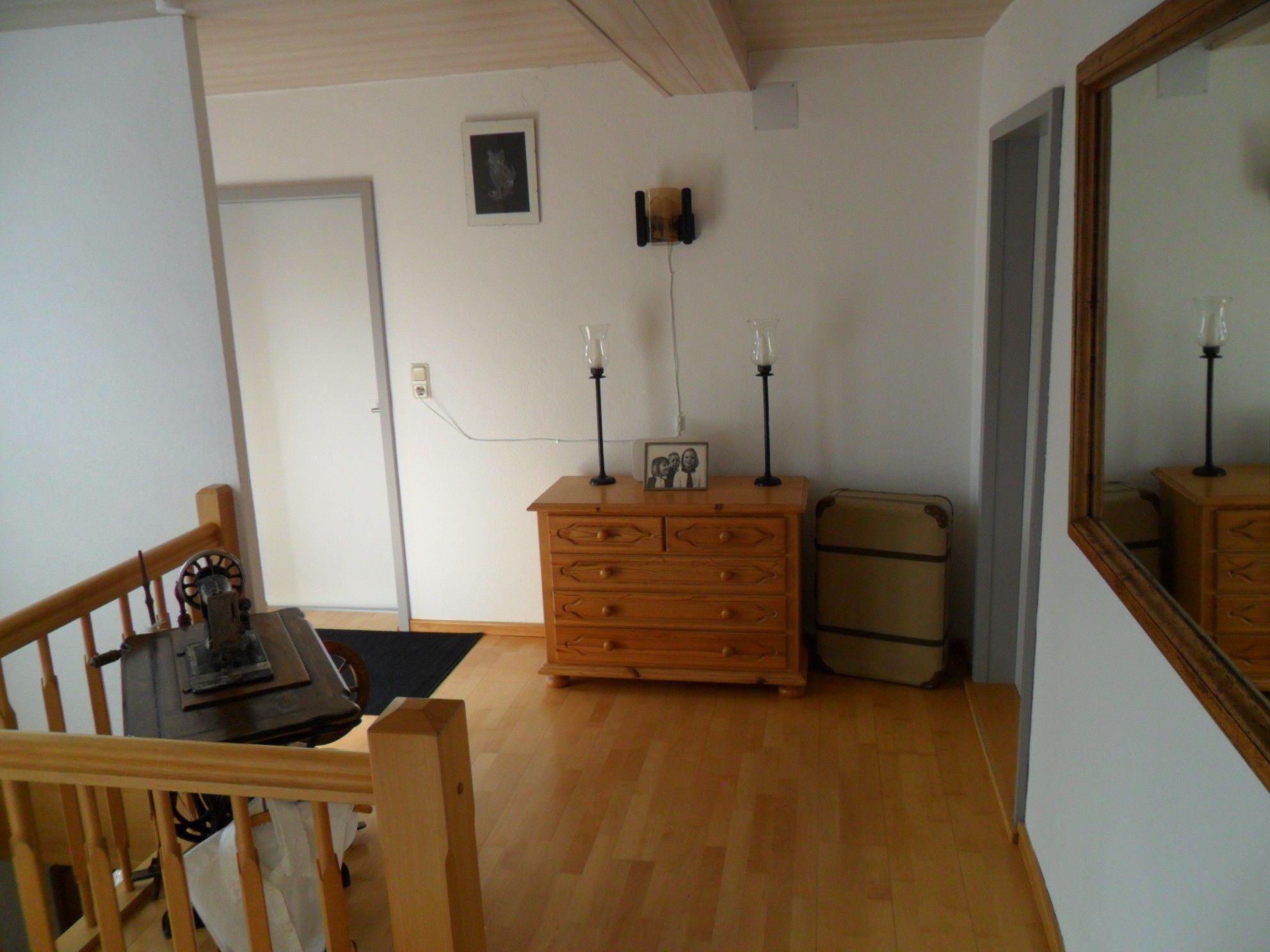 Ein Zimmer mit einem hölzernen Schminktisch, einem Koffer, einem Tisch mit Stuhl und einem Spiegel an der Wand.