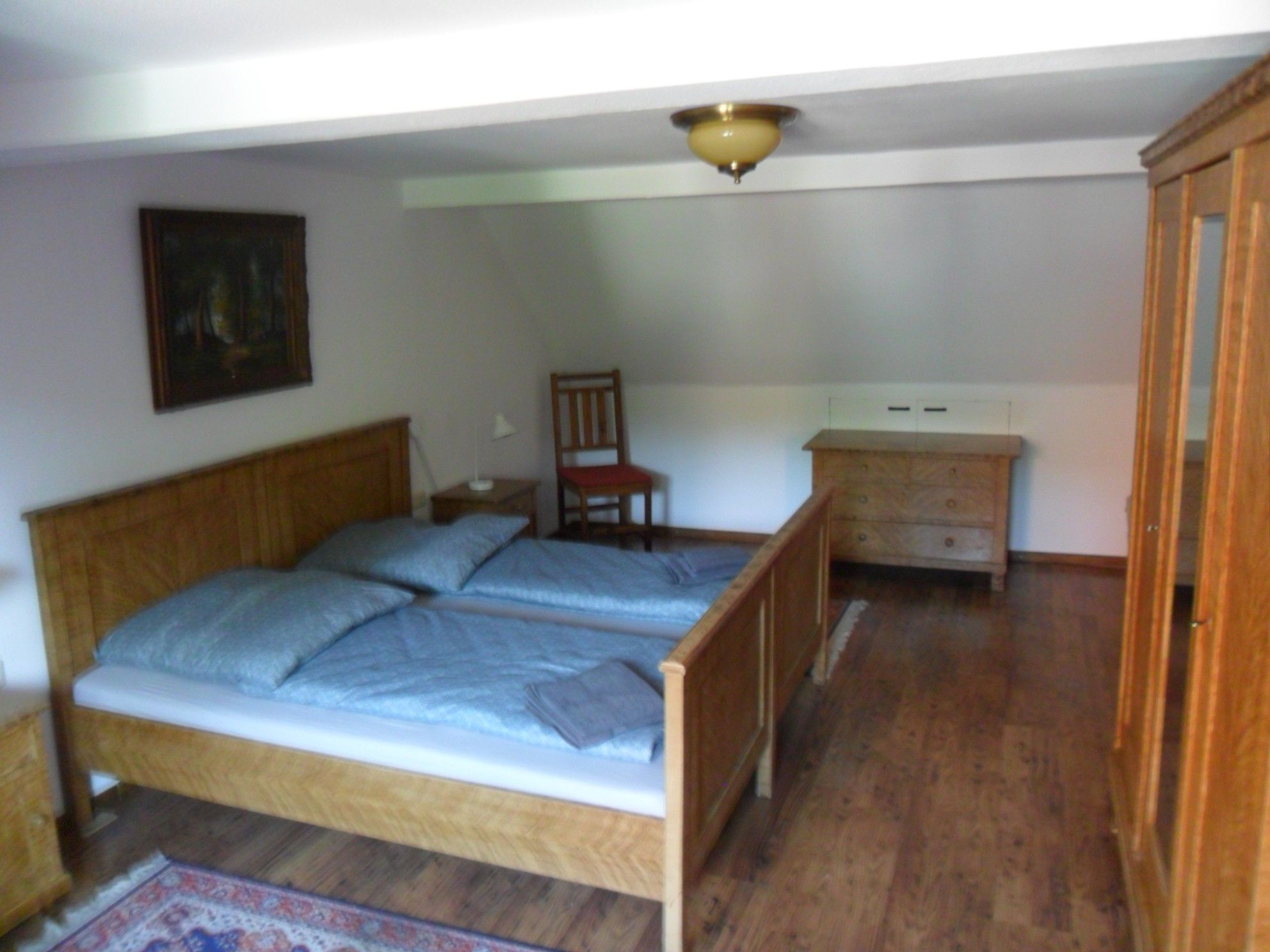 Ein Schlafzimmer mit einem hölzernen Bett, einem Sessel, einem Nachttisch, einer Kommode und einer Deckenleuchte.