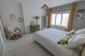 Un dormitori amb llit, cadira, tocador, planta i finestra amb cortines.