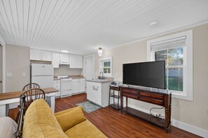Una cocina compacta con armarios blancos, estufa y lavamanos. Hay un comedor con una mesa y sillas de madera, y un televisor sobre un mueble de madera.