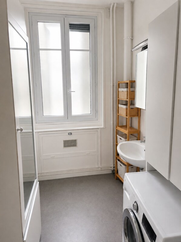 Une salle de bain avec une fenêtre, un lavabo, un miroir, un lave-linge et une étagère en bois.