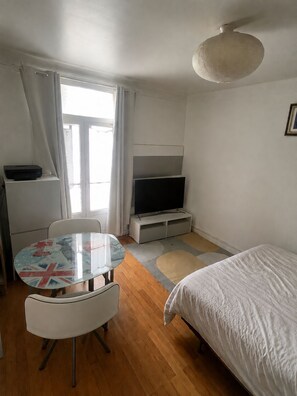 Une pièce comprenant un lit, un téléviseur posé sur un meuble, une table avec des chaises et une fenêtre avec des rideaux.