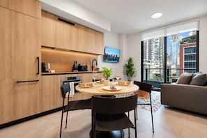 A modern kitchen with wooden cabinets, a dining area with a round table, and a living space with a sofa and large windows overlooking a cityscape. - NEW   High-Rise 1BR Den  Rooftop Pool   Gym 402 (Miami)