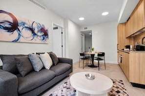 A modern living room with a grey sofa, a dining table with chairs, and a kitchen area with wooden cabinets. - NEW   High-Rise 1BR Den  Rooftop Pool   Gym 402 (Miami)