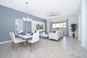 A modern living room with a dining area, a ceiling fan, and a large window with curtains. - NEW   2Bd Retreat with Game Room   Lazy River 9333 (Davenport)