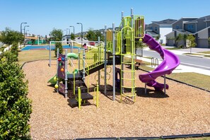 A playground with a purple slide and climbing structures, surrounded by a landscaped area with mulch and small trees. - NEW   2Bd Retreat with Game Room   Lazy River 9333 (Davenport)