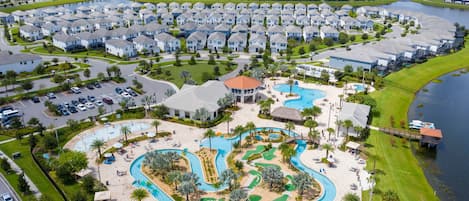 A residential area with a large swimming pool complex, including a main pool and a water slide, surrounded by houses and a body of water.