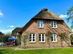 Ein Backsteinhaus mit Strohdach, zahlreichen Fenstern und einem kleinen Garten mit Tisch und Stühlen.